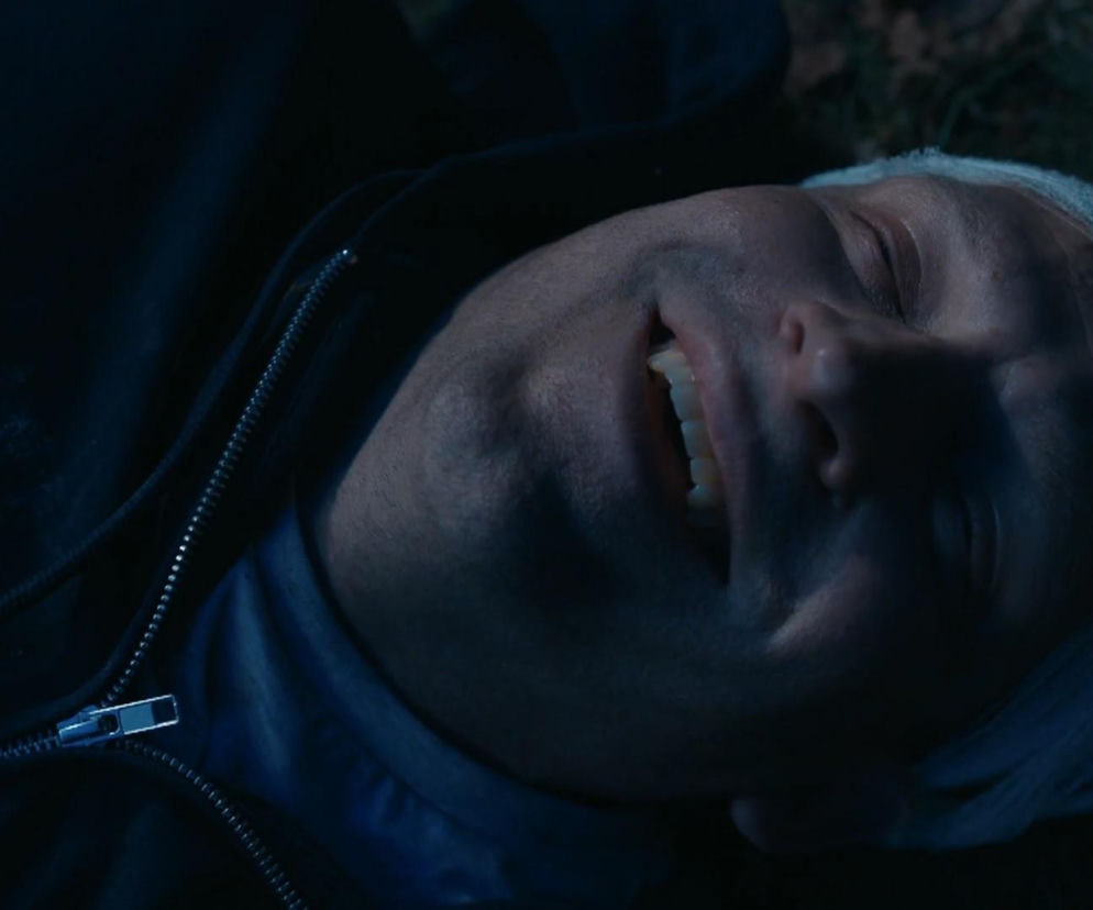 Mężczyzna z zamkniętymi oczami i otwartymi ustami, wyrażający ulgę i radość, leży na ziemi w ciemności. Na głowie ma widoczne ślady taśmy, a na sobie ciemną kurtkę z rozpiętym suwakiem, co nawiązuje do ucieczki Łukasza z więzienia Agnieszki w serialu Barwy szczęścia, o czym można przeczytać na portalu Super Seriale.