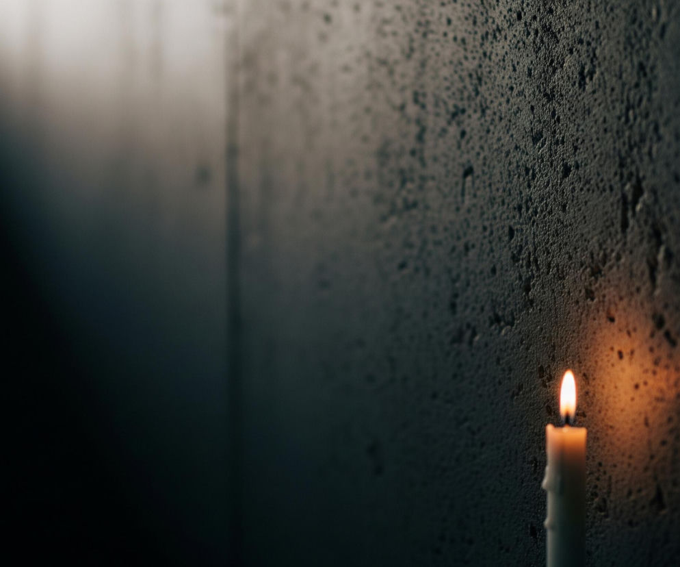 W dolnej prawej części obrazu znajduje się zapalona, biała świeca z pomarańczowo-żółtym płomieniem. Świeca jest częściowo stopiona, z widocznymi spływami wosku po jej bokach. Tło stanowi ciemna, chropowata ściana w odcieniach szarości i brązu, z wyraźną teksturą złożoną z wielu drobnych nierówności i wgłębień. Górny lewy róg obrazu rozświetla skośny, jasny pas światła, przechodzący w ciemniejszy obszar.