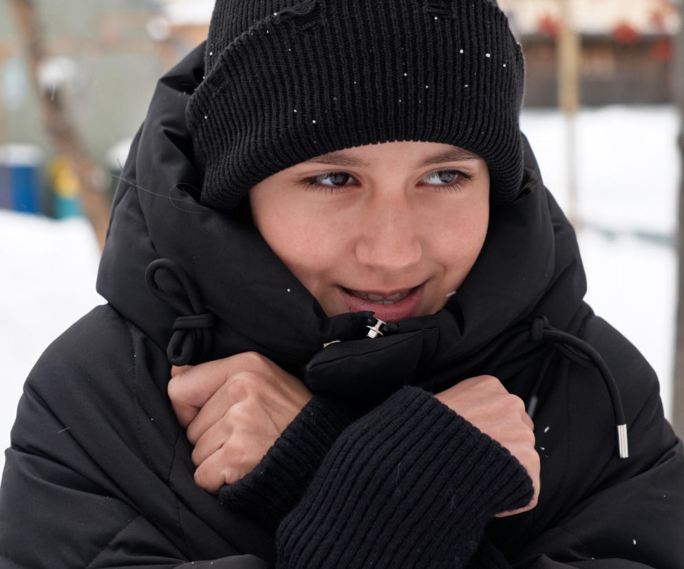 Młoda osoba w czarnej puchowej kurtce i wełnianej czapce, obejmująca się dłońmi, by się ogrzać. Na tle zaśnieżonego otoczenia widać opady śniegu, co podkreśla mroźną pogodę, o której pisze Poradnik Zdrowie.