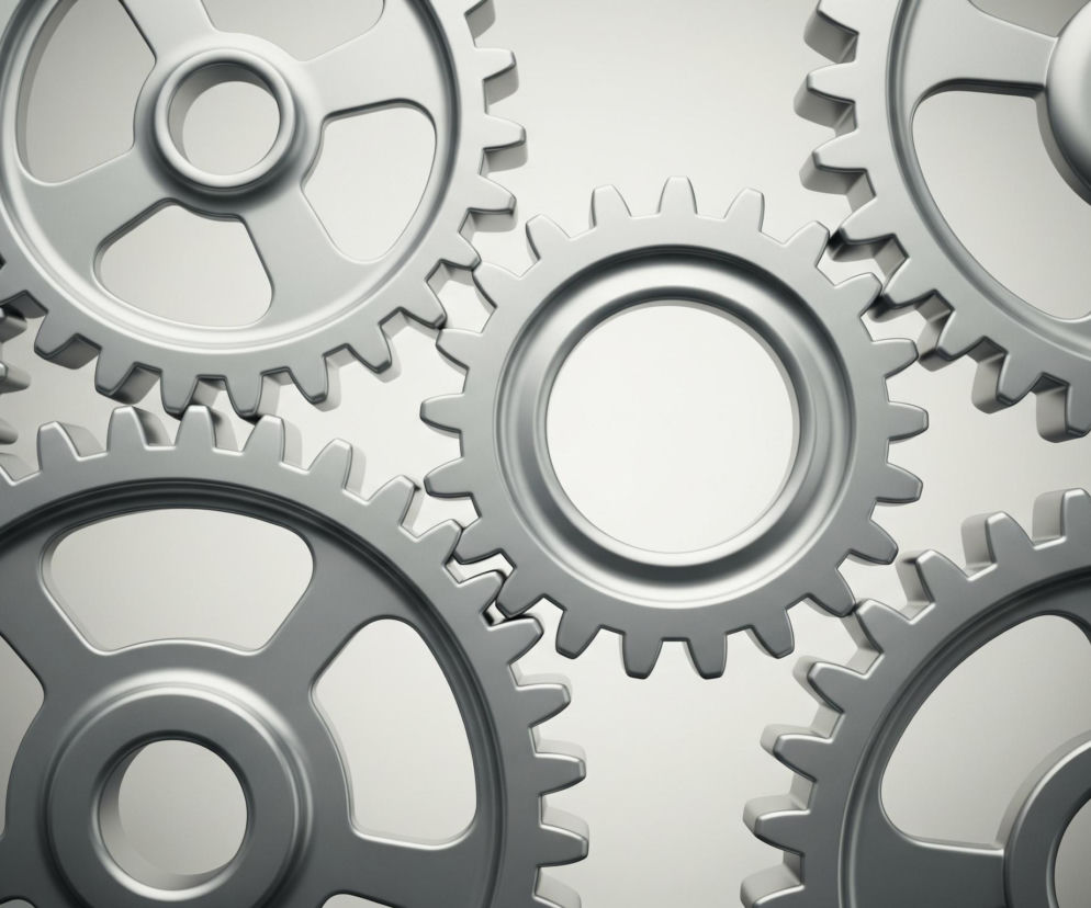 Skupisko siedmiu kół zębatych o metalicznym, srebrzystym odcieniu, które są ze sobą zazębione i tworzą spójną całość mechanizmu. Koła te mają różne rozmiary, od mniejszych, znajdujących się głównie po lewej stronie, po większe, dominujące w centralnej i prawej części kompozycji. Wszystkie koła posiadają w centralnej części otwory o obrysie okręgu, a niektóre z nich mają również dodatkowe wycięcia w ramionach, nadające im ażurowy wygląd. Całość jest osadzona na jasnym, neutralnym tle, które płynnie przechodzi od bieli do jasnej szarości, a subtelne cienie i refleksy świetlne podkreślają trójwymiarowość kół.