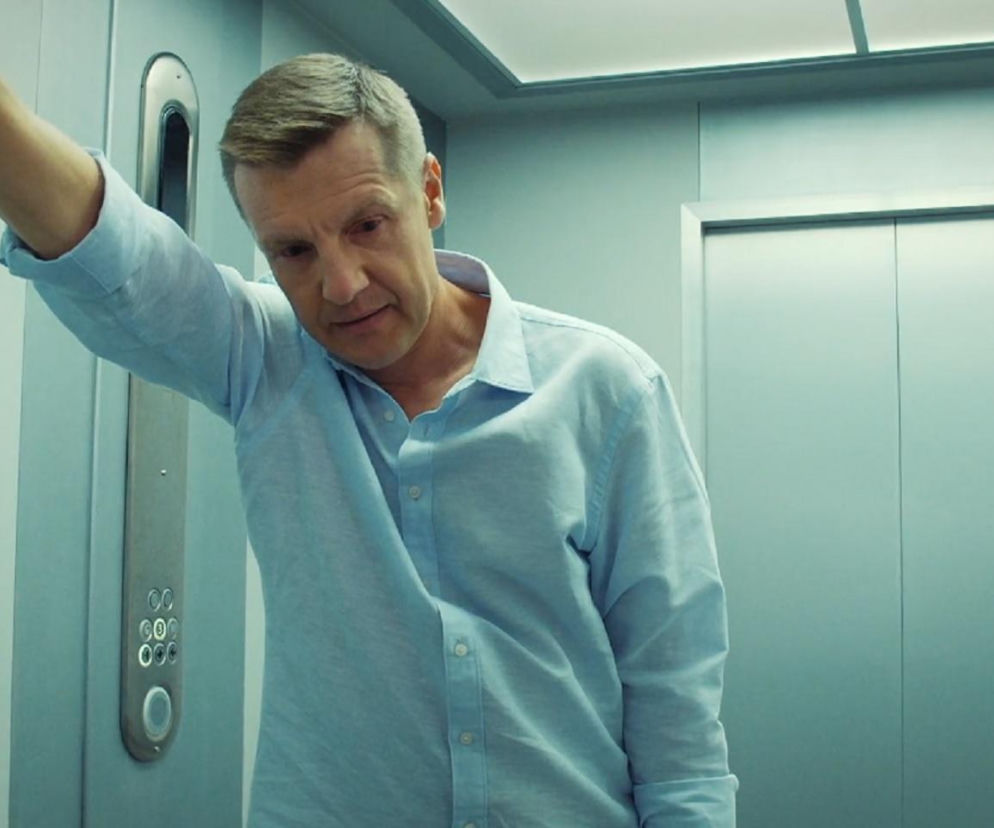 Paweł (Rafał Mroczek) w jasnoniebieskiej koszuli opiera się o ścianę windy, patrząc w dół z zatroskaną miną. Widok ten ilustruje jego zmagania po śmierci Franki, kiedy to Antoś zacznie przeszkadzać mu w pracy. Więcej o dramatycznych losach Pawła znajdziesz na Super Seriale.