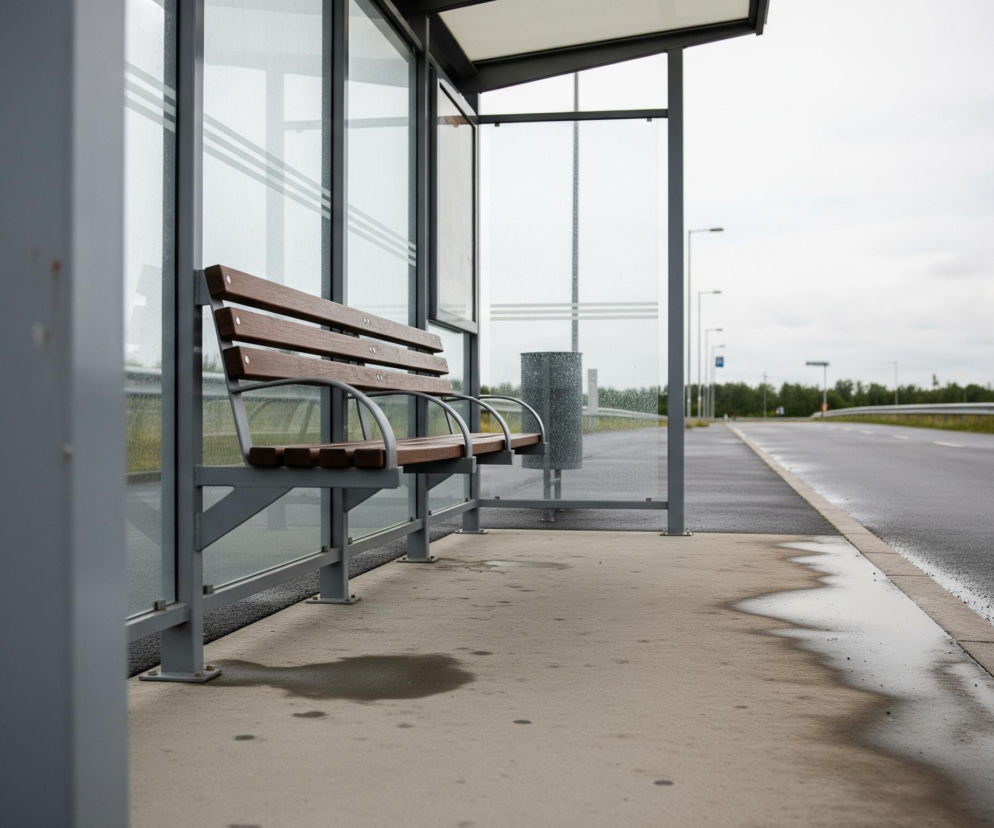 Przystanek autobusowy z ławką, oszklonymi ścianami i zadaszeniem. Na betonowej podłodze widać mokre plamy, a na szybach krople deszczu. Po prawej stronie, obok przystanku, biegnie asfaltowa droga z białym oznakowaniem, a w oddali widać słupy oświetleniowe i fragmenty zieleni pod szarym, zachmurzonym niebem.
