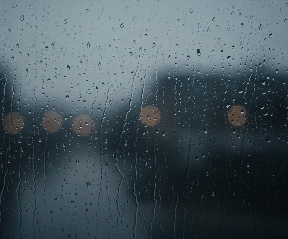 Przednia szyba samochodu pokryta jest licznymi kroplami deszczu, niektóre z nich spływają smugami pionowo w dół. Na tle szyby widać rozmazane, nieostre kształty budynków w szarych odcieniach, a poniżej nich pięć okrągłych, bursztynowych świateł, równomiernie rozmieszczonych poziomo. Cały obraz utrzymany jest w ciemnej, chłodnej tonacji szarości i granatu, co potęguje wrażenie deszczowego dnia lub zmierzchu.
