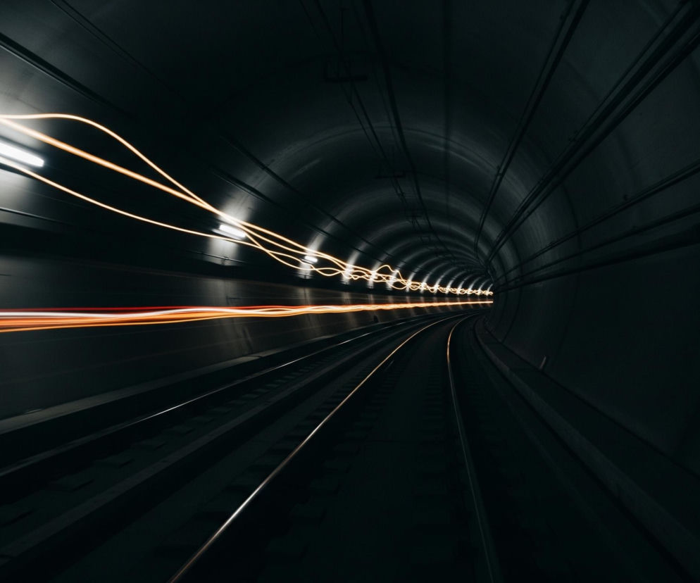 Długi tunel z torami kolejowymi rozciąga się w głąb obrazu, zakręcając w prawo w oddali. Na ciemnych ścianach tunelu i suficie widoczne są ślady smugi światła o barwie pomarańczowo-żółtej, biegnące z lewej strony kadru wzdłuż góry i dolnej części tunelu, świadczące o ruchu. Dwa metalowe tory biegnące przez tunel odbijają światło, tworząc jasne, cienkie linie na ciemnym tle. Całość jest skąpana w mrocznym, niemal czarnym oświetleniu z akcentami jaśniejszych smug światła.