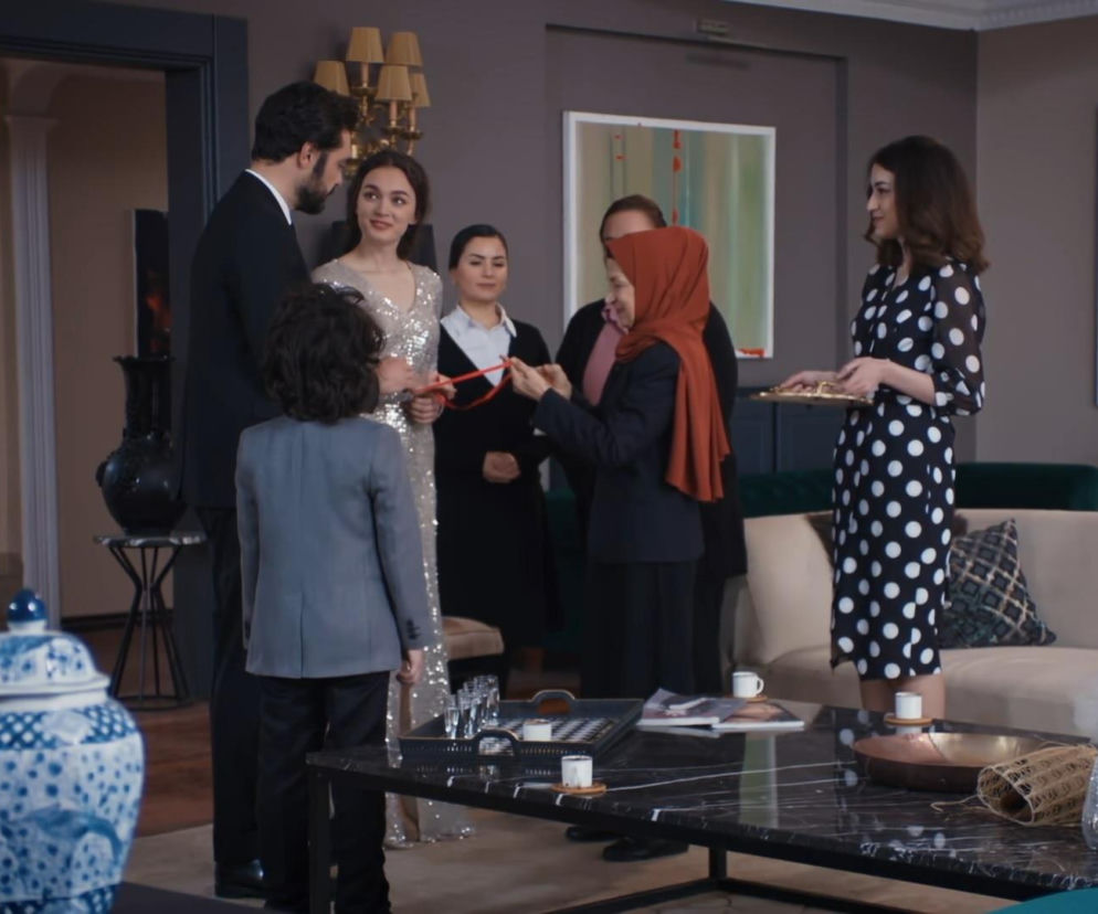 Yaman i Nana, ubrana w błyszczącą sukienkę, podczas oficjalnych zaręczyn w salonie, w otoczeniu bliskich i służby. Nana trzyma za rękę chłopca w szarym garniturze, a inna kobieta w hidżabie trzyma czerwone wstążki do obrączek. Kobieta w sukience w groszki podaje tacę z kawą, o czym przeczytasz na Super Seriale.