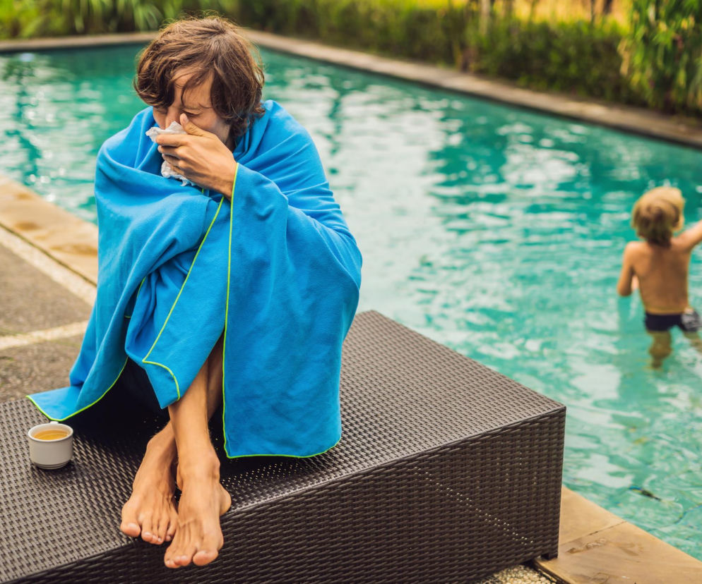 Kobieta z objawami przeziębienia, owinięta niebieskim ręcznikiem, siedzi przy basenie z kubkiem herbaty, podczas gdy dziecko bawi się w wodzie. Na Poradnik Zdrowie przeczytasz o chorobach na urlopie i tym, jak przygotować organizm przed wyjazdem.