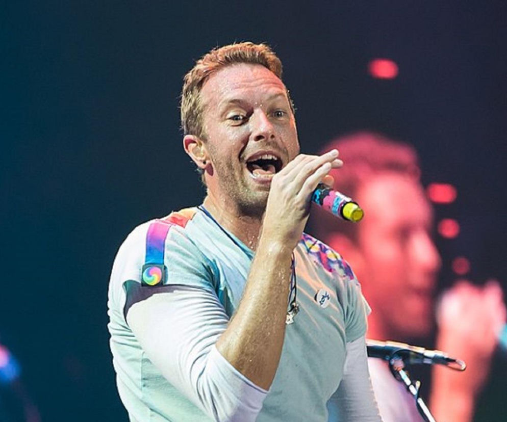 Chris Martin śpiewa do kolorowego mikrofonu podczas koncertu. O karierze lidera Coldplay przeczytasz na EskaRock.