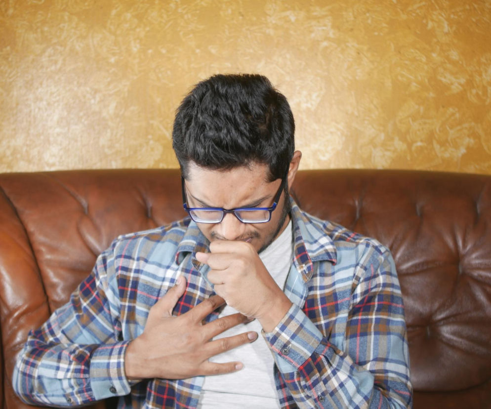 Mężczyzna w okularach i koszuli w kratę siedzi na skórzanej kanapie, zakrywając usta dłonią i trzymając drugą na klatce piersiowej, co symbolizuje czkawkę lub dolegliwości oddechowe. Przeczytaj na Poradnik Zdrowie, jak radzić sobie z tym problemem.
