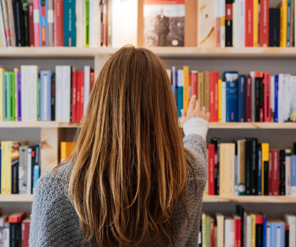 Kobieta z długimi brązowymi włosami, ubrana w szary sweter, sięga po książkę z drewnianej półki wypełnionej kolorowymi woluminami. Scena symbolizuje relaks, rozwijanie koncentracji i wzmacnianie empatii, o czym więcej przeczytasz na Poradnik Zdrowie.