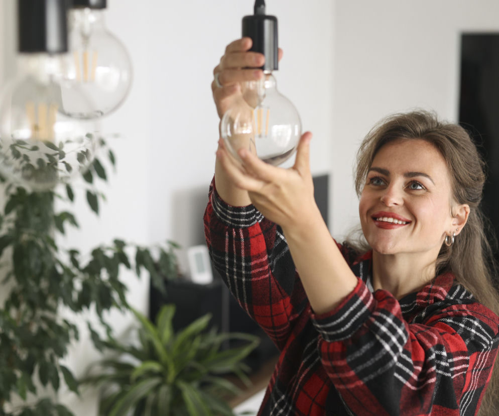 Uśmiechnięta kobieta w koszuli w kratę wkręca żarówkę. O oszczędzaniu energii w domu przeczytasz na Muratordom.