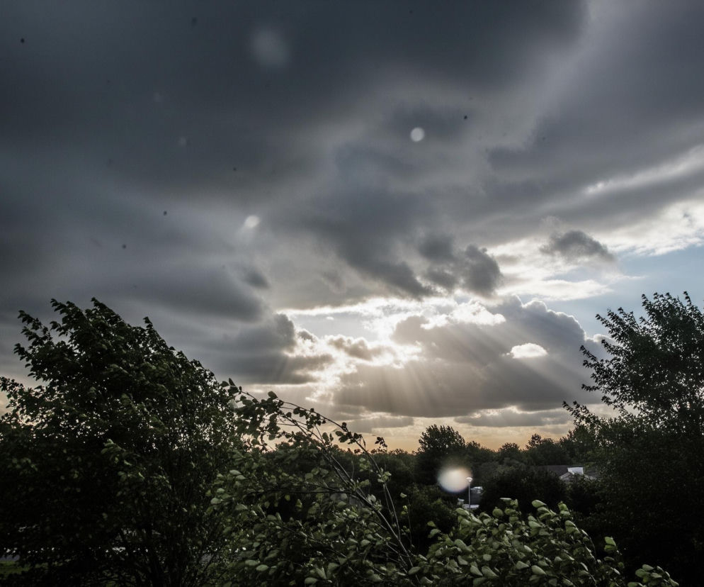 Niebo dominuje w górnej części obrazu, ukazując dramatyczne ciemne chmury, częściowo zasłaniające słońce, z którego przebijają się jasne promienie światła w centrum. Poniżej rozciąga się krajobraz z ciemnozielonymi drzewami i krzewami w pierwszym planie, a w oddali widać fragmenty zabudowy, w tym biały dom po lewej stronie. Na zdjęciu widoczne są liczne jasne, okrągłe artefakty, przypominające odbicia światła lub krople wody, oraz mniejsze, ciemne plamki, rozproszone po całej kompozycji.