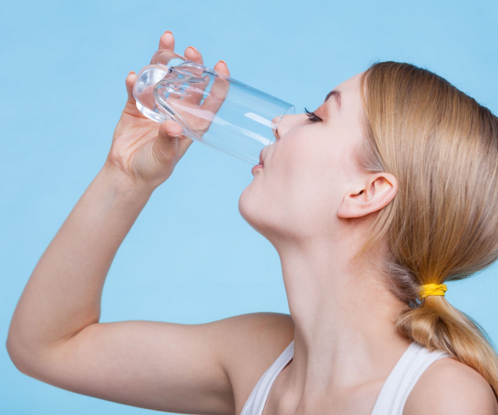 Młoda blondynka w białej koszulce pije wodę z przezroczystej szklanki na jasnoniebieskim tle, ukazując nawadnianie i zdrowy styl życia. Więcej o wodzie w kranie i jej wpływie na zdrowie dowiesz się na Poradnik Zdrowie.