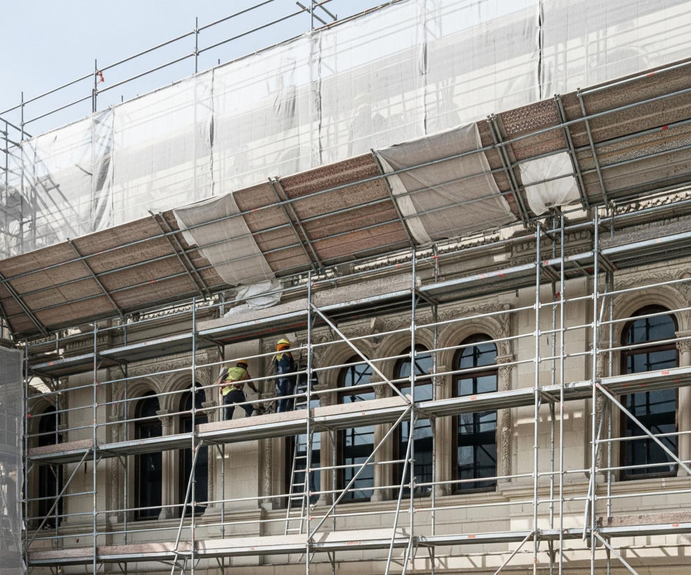 Srebrne rusztowania, częściowo pokryte jasną, białą siatką, otaczają górną część jasnego budynku o skomplikowanej architekturze. Na jednym z poziomów rusztowania widoczni są dwaj robotnicy w kaskach ochronnych i odzieży roboczej, jeden z nich ma żółtą kamizelkę i kask, drugi pomarańczowy. Tło stanowi błękitne niebo z kilkoma chmurami, a w szczelinach między rusztowaniami widoczne są ozdobne detale budynku i ciemne, półokrągłe okna.