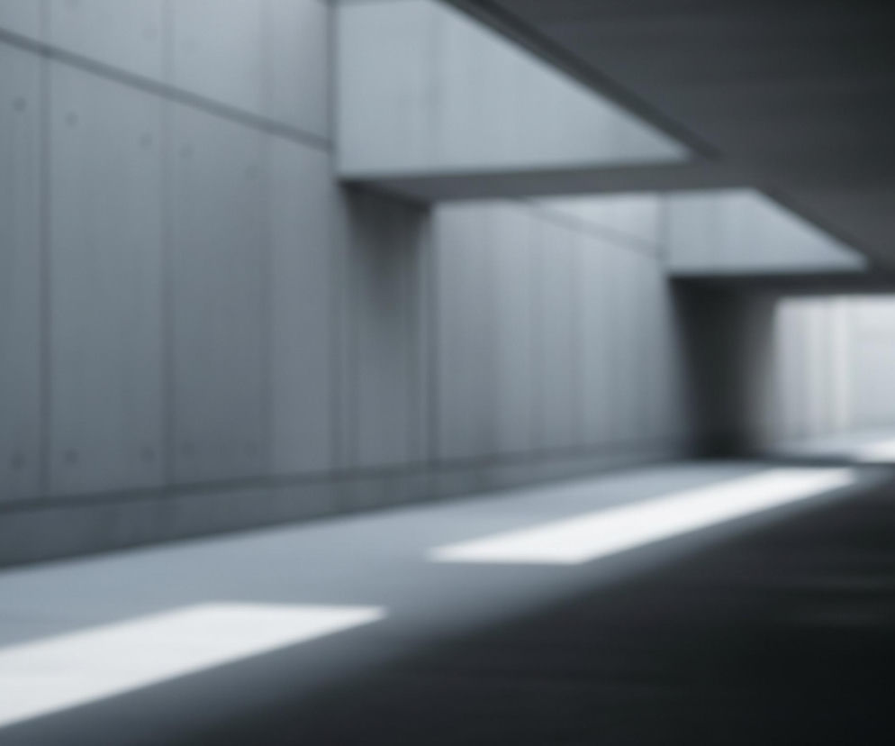 Wnętrze długiego, pustego korytarza, wykonanego z betonu w odcieniach szarości. Światło wpada przez otwory w suficie i z boku, tworząc jasne prostokątne plamy na podłodze i ścianie. Dalej korytarz staje się ciemniejszy i niknie w oddali.