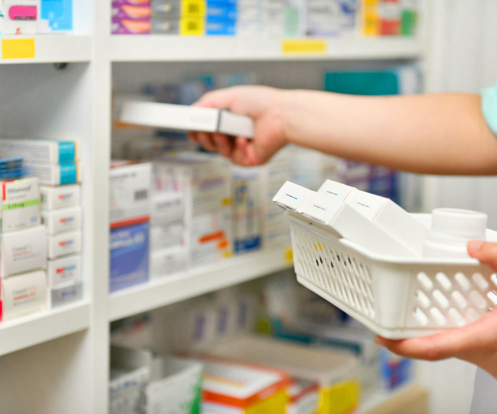 Farmaceuta układa opakowania leków na półkach w aptece. Osoba w białym fartuchu wybiera i przenosi leki do koszyka, co symbolizuje nową możliwość realizowania e-recept w różnych aptekach, o czym można przeczytać na Super Biznes.