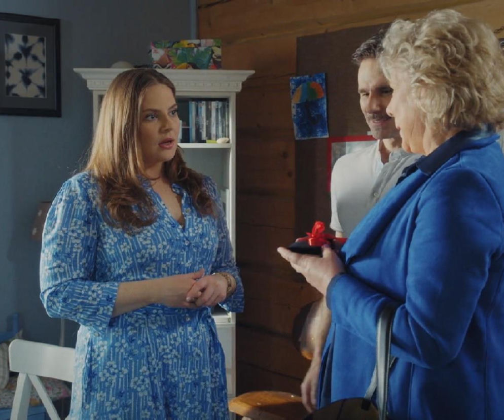 Melania Grzesiewicz (Anita) w niebieskiej sukience, Marcin Bosak (Kamil) w białej koszulce i Dorota Kamińska (Anna) w niebieskim płaszczu rozmawiają w mieszkaniu. Anna trzyma czarne pudełko z czerwoną kokardką. Więcej o ich losach w M jak miłość na Super Seriale.