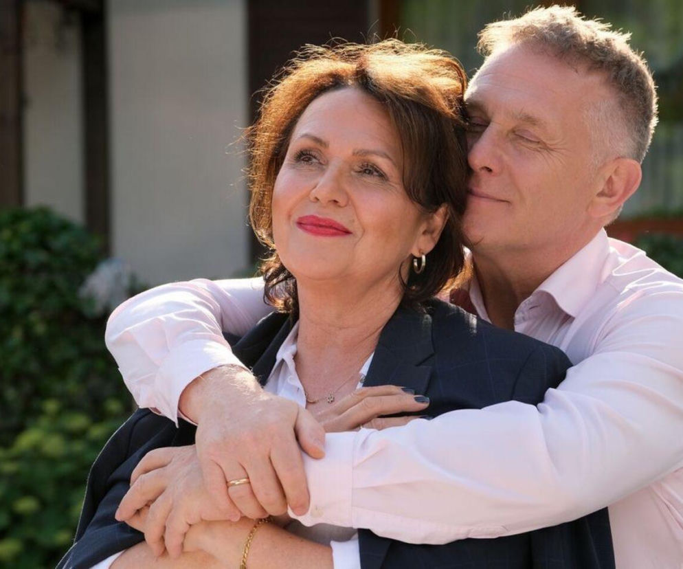 Uśmiechnięta kobieta z brązowymi włosami i czerwonymi ustami, w białej koszuli i granatowej marynarce, jest obejmowana od tyłu przez mężczyznę w jasnoróżowej koszuli, z zamkniętymi oczami. To kadr z serialu M jak miłość, o którego przerwie w emisji przeczytasz na stronie Super Seriale.