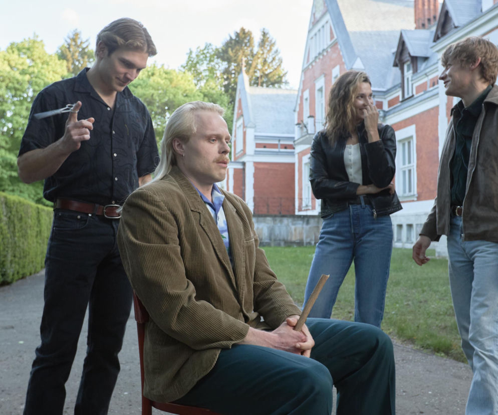 Grupa młodych agentów z serialu Wojna zastępcza podczas szkolenia w Kiejkutach. Waleria Gorobets (Zuza), Piotr Witkowski (Budyń), Karol Bernacki (Gutek) i Mateusz Więcławek (Artur) pozują przed budynkiem szkoły, przygotowując się do misji. Więcej o ich losach na stronie Super Seriale.