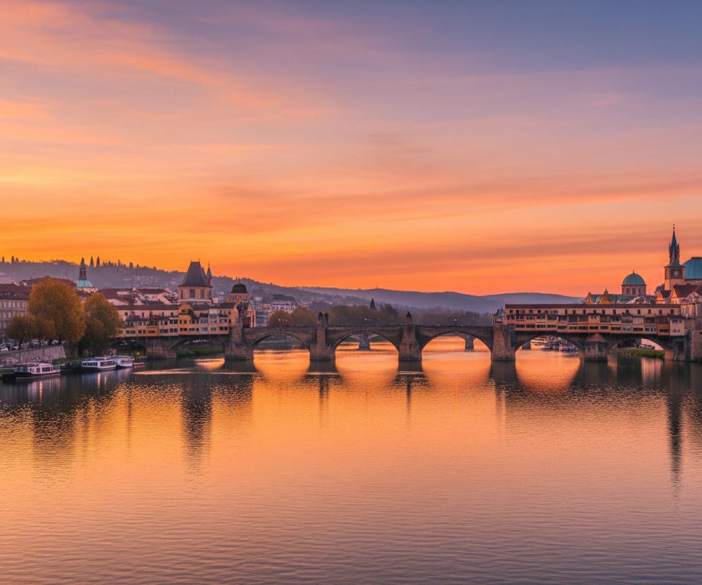 Szeroki kadr ukazuje panoramę miasta rozciągającą się wzdłuż rzeki, oświetloną ciepłym światłem zachodzącego słońca. Dominującym elementem jest kamienny most z kilkoma arkadami, częściowo zabudowany budynkami z beżowymi i brązowymi elewacjami oraz pomarańczowymi dachami, połączony z długim, niższym mostem z drewnianymi konstrukcjami. Po obu stronach rzeki widać rzędy różnokolorowych kamienic z licznymi oknami, niektóre z nich posiadają zielone kopuły i smukłe wieże z zegarami. Całość krajobrazu odbija się w spokojnych wodach rzeki, tworząc pomarańczowo-różowe refleksy, a niebo nad miastem mieni się odcieniami od jaskrawej pomarańczy przy horyzoncie po delikatny błękit w górnej części kadru.