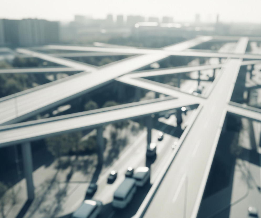 Rozmyty obraz przedstawia z lotu ptaka skrzyżowanie autostrad z kilkoma poziomami estakad, przecinających się pod różnymi kątami. Dominują tu szare odcienie betonu, z elementami zieleni drzew i krzewów widocznych między konstrukcjami. W oddali majaczy niewyraźny zarys miejskiej panoramy z licznymi budynkami. Na niższych poziomach dróg widać kilka rozmazanych samochodów i autobusów, poruszających się w różnych kierunkach.