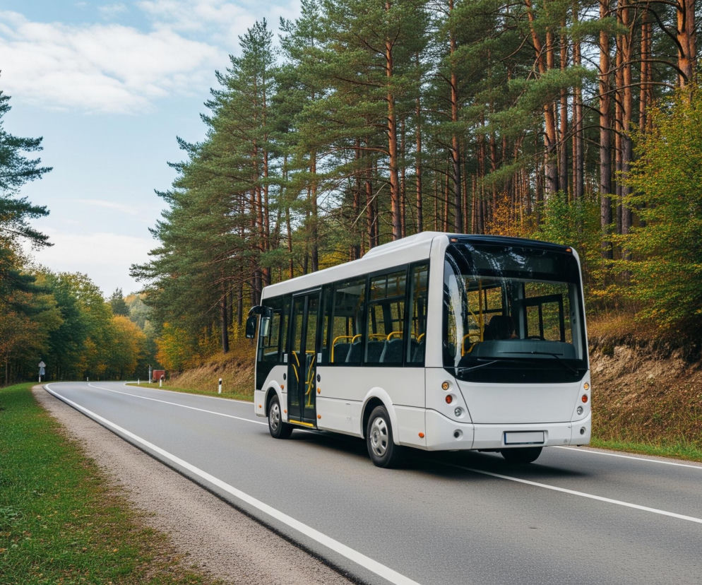 Biały autobus elektryczny z otwartymi drzwiami stoi na asfaltowej drodze, biegnącej przez las z przewagą sosen. Po bokach drogi zielone pobocza. Zdjęcie ilustruje temat transportu ekologicznego w kontekście artykułu na Super Biznes.