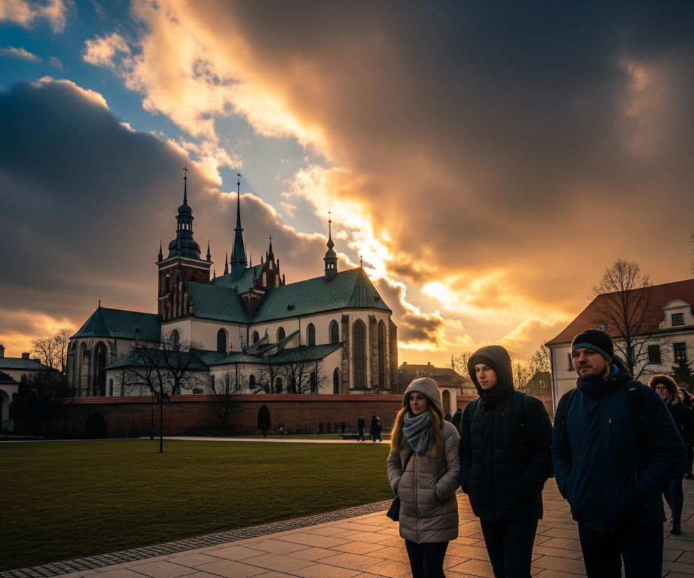 Trzy osoby, dwie kobiety i jeden mężczyzna, w zimowych kurtkach i czapkach idą po chodniku po prawej stronie kadru, zwrócone lekko w prawo od obserwatora. Po lewej stronie dominuje duży, zabytkowy budynek z zielonym dachem i wieloma wieżyczkami z krzyżami, otoczony murem z czerwonej cegły i zielonym trawnikiem. Tło stanowi dramatyczne niebo z ciemnymi chmurami na górze i jasnym, pomarańczowo-złotym światłem zachodzącego słońca przebijającym się przez chmury po prawej stronie i w centrum. W oddali, za ludźmi, widoczne są inne budynki z czerwonymi dachami oraz drzewa bez liści.