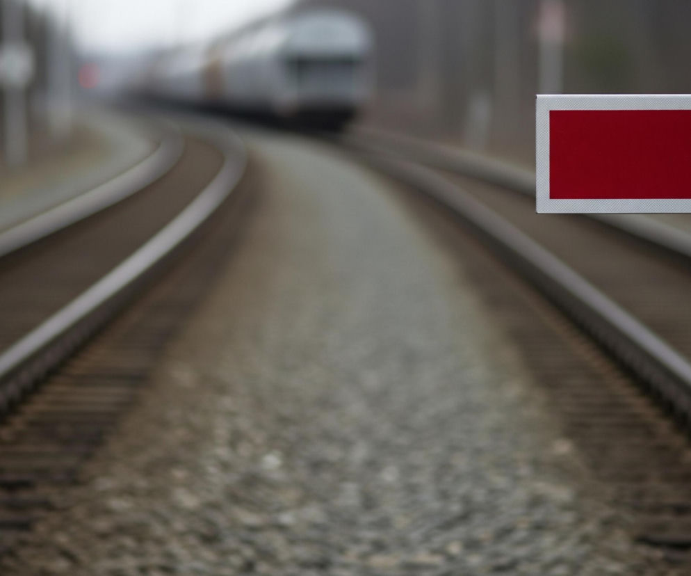 Płaski, prostokątny, srebrno-szary przedmiot z widoczną wklęsłą, czerwoną powierzchnią zajmuje prawą część kadru, częściowo wychodząc poza jego ramy. Ostry obiekt z drobnym, ziarnistym wzorem na powierzchni jest ustawiony prostopadle do linii kolejowych, które dominują w centrum kadru, rozciągając się od dołu w kierunku góry i zbiegając się w dali. Szyny kolejowe leżą na podsypce żwirowej, która stopniowo staje się niewyraźna, podobnie jak oddalający się pociąg o jasnych barwach, majaczący w tle.
