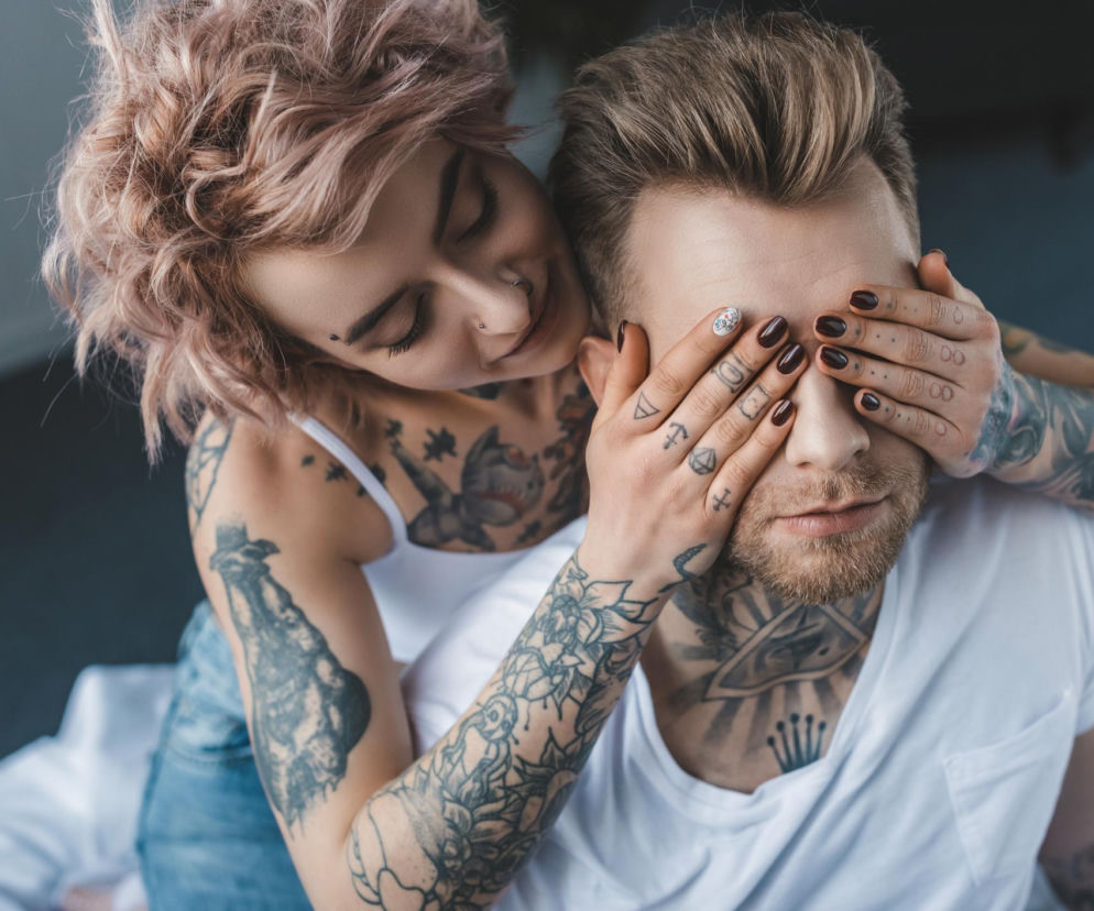 Para z tatuażami, kobieta zakrywająca oczy mężczyźnie. Oboje mają liczne tatuaże, m.in. na rękach, szyi i klatce piersiowej. Zdjęcie ilustruje temat wpływu tatuaży na układ odpornościowy, o którym można przeczytać na Poradniku Zdrowie.