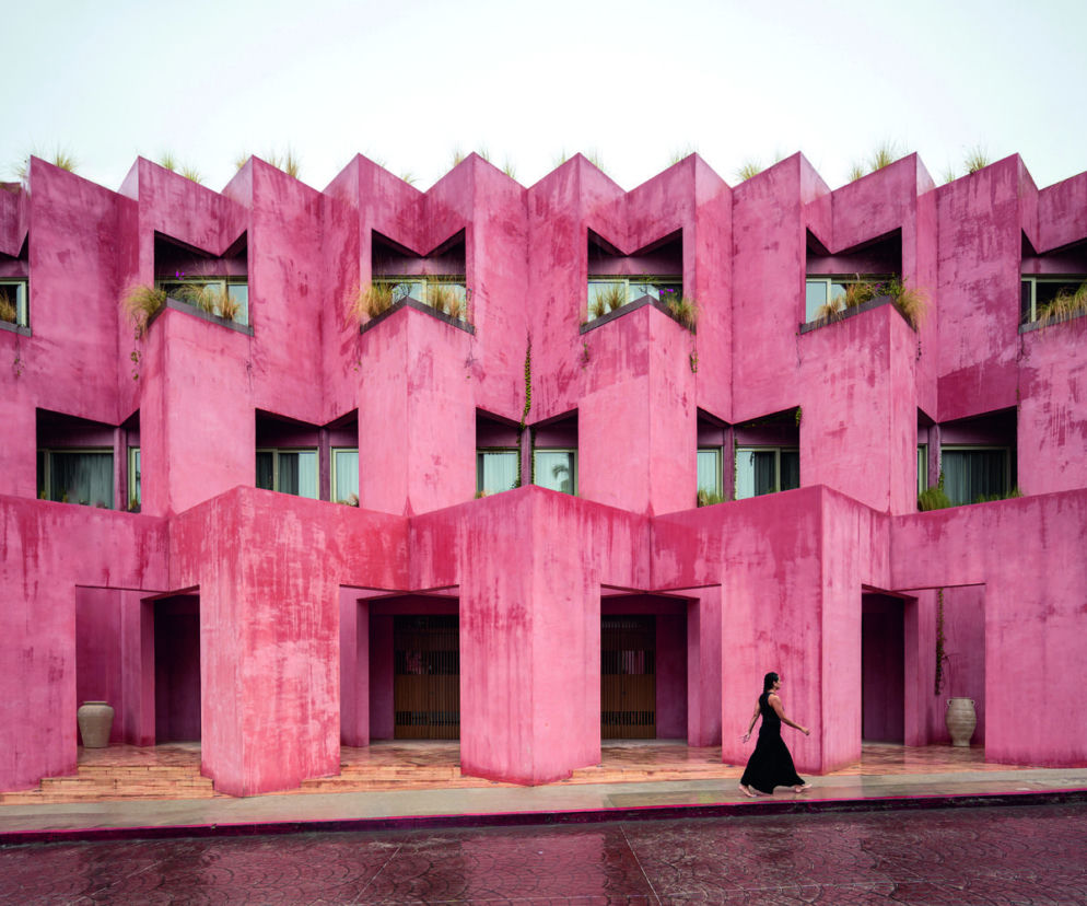 Różowy, geometryczny budynek hotelu Laiva Plaza w San José del Cabo, Meksyku, z zielenią na dachach i widoczną kobietą idącą chodnikiem. Architektura Murator Plus poleca ten obiekt, który wyróżnia się nietypową elewacją inspirowaną meksykańskimi papel picado.