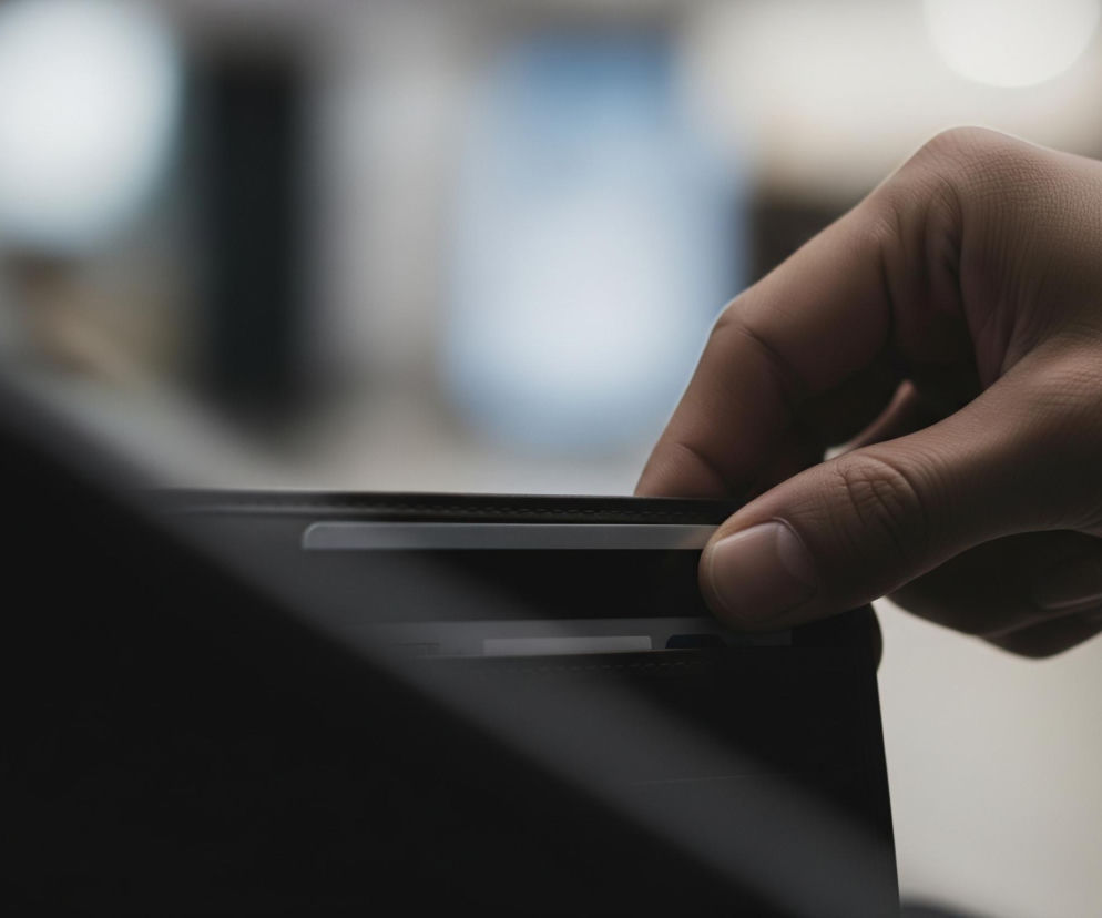 Prawa dłoń o ciemniejszej karnacji, z palcami częściowo zgiętymi, wyjmuje białą kartę z ciemnobrązowego portfela. Paznokieć kciuka jest wyraźnie widoczny, naciskający na krawędź karty. Górna część portfela, wykonana z materiału przypominającego skórę, jest widoczna, z drobnymi przeszyciami biegnącymi wzdłuż krawędzi. W tle, poza ostrością, widoczne są rozmyte plamy światła w odcieniach bieli i jasnego błękitu.