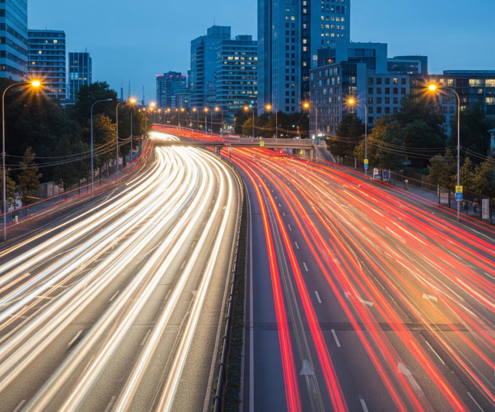 Długa ekspozycja ukazuje dynamiczny ruch uliczny na wielopasmowej drodze w nocy lub o zmierzchu. Po lewej stronie jezdni widoczne są białe smugi świateł pojazdów jadących w kierunku oddalającym się od widza, natomiast po prawej stronie dominują czerwone smugi świateł tylnych pojazdów jadących w kierunku widza. Droga rozciąga się przez centrum kadru, a po jej obu stronach widać drzewa, latarnie uliczne emitujące ciepłe, żółte światło, oraz rzędy wysokich budynków z licznymi oknami, w których świecą światła. Tło stanowią liczne, ciemne drapacze chmur i niższe budynki, oświetlone przez niebo w odcieniach głębokiego błękitu.