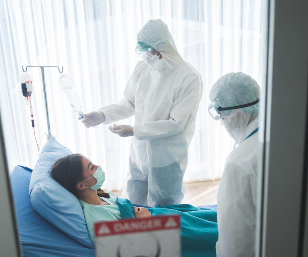 Pacjentka z maseczką leży w szpitalnym łóżku, a dwóch pracowników medycznych w kombinezonach ochronnych i maseczkach zajmuje się kroplówką. Widoczna tabliczka ostrzegawcza Danger podkreśla sytuację epidemiczną. O ograniczeniach w odwiedzinach przeczytasz na Poradnik Zdrowie.