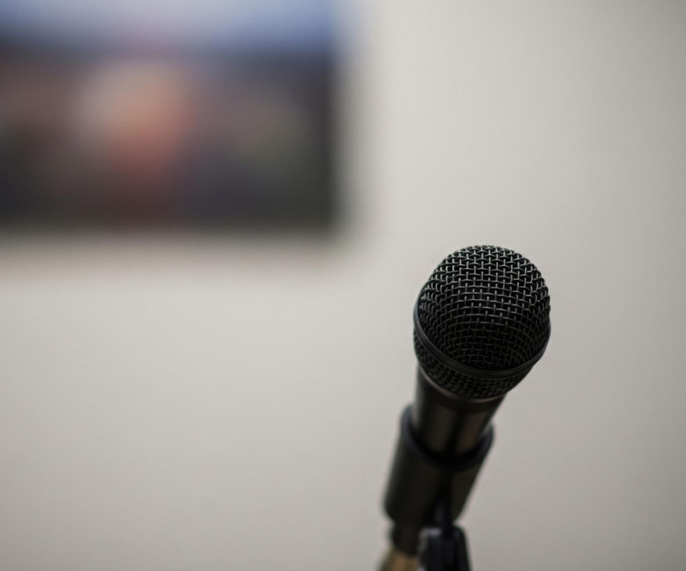 Ciemny mikrofon studyjny z metalową siatką na górze znajduje się w centrum kadru, lekko nachylony w prawą stronę. Stoi na czarnym statywie, który jest częściowo widoczny u dołu. Tło stanowi jasna, beżowa ściana, a w górnym lewym rogu widać rozmazany, prostokątny kształt z niewyraźnymi odcieniami niebieskiego, brązu i czerni, prawdopodobnie ekran lub obraz.