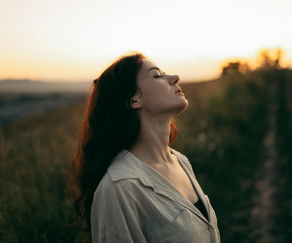 Młoda kobieta z zamkniętymi oczami unosi twarz do góry, oddychając spokojnie w tle zachodzącego słońca, symbolizując relaks i techniki oddechowe. Na Poradnik Zdrowie można przeczytać, jak świadome oddychanie wpływa na obniżenie ciśnienia krwi i wyciszenie układu nerwowego.