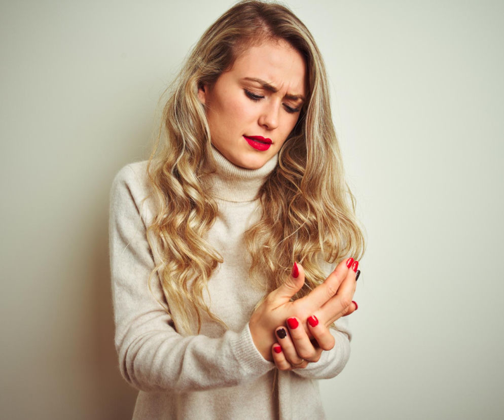 Kobieta z blond włosami, w beżowym golfie, trzymająca dłonie, z czerwoną szminką i pomalowanymi paznokciami, odczuwająca ból lub niepokój związany z elektryzowaniem się. To sygnał alarmowy organizmu, o którym przeczytasz więcej na Poradnik Zdrowie.