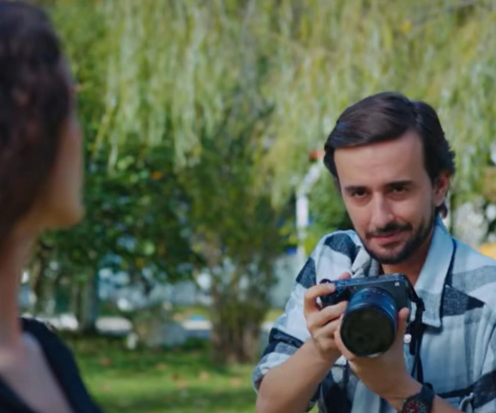 Mężczyzna Semih w koszuli w biało-czarną kratę, trzymający aparat fotograficzny z obiektywem, patrzy w kierunku kobiety Nany, która odwraca się do niego plecami. Ta scena z serialu Dziedzictwo może prowadzić do tragicznych konsekwencji, jak przeczytasz na Super Seriale.
