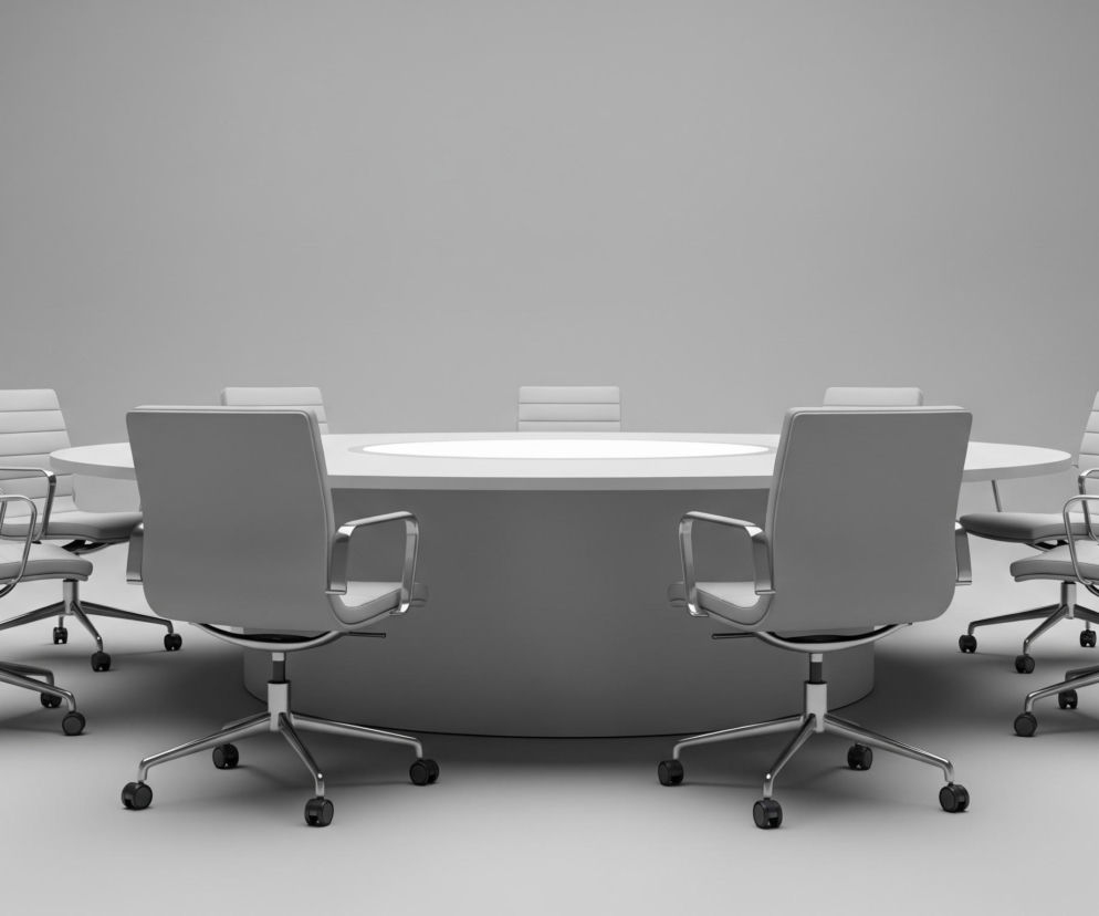 W centrum znajduje się okrągły stół konferencyjny w kolorze jasnoszarym, z jaśniejszą, niemal białą powierzchnią blatu. Wokół stołu rozmieszczono osiem identycznych, obrotowych krzeseł biurowych z kółkami, utrzymanych w tym samym odcieniu szarości. Krzesła mają chromowane metalowe ramy, podłokietniki oraz podstawy w kształcie gwiazdy z pięcioma ramionami. Tło stanowi jednolita, gładka ściana i podłoga w kolorze szarym, tworząc minimalistyczną przestrzeń.