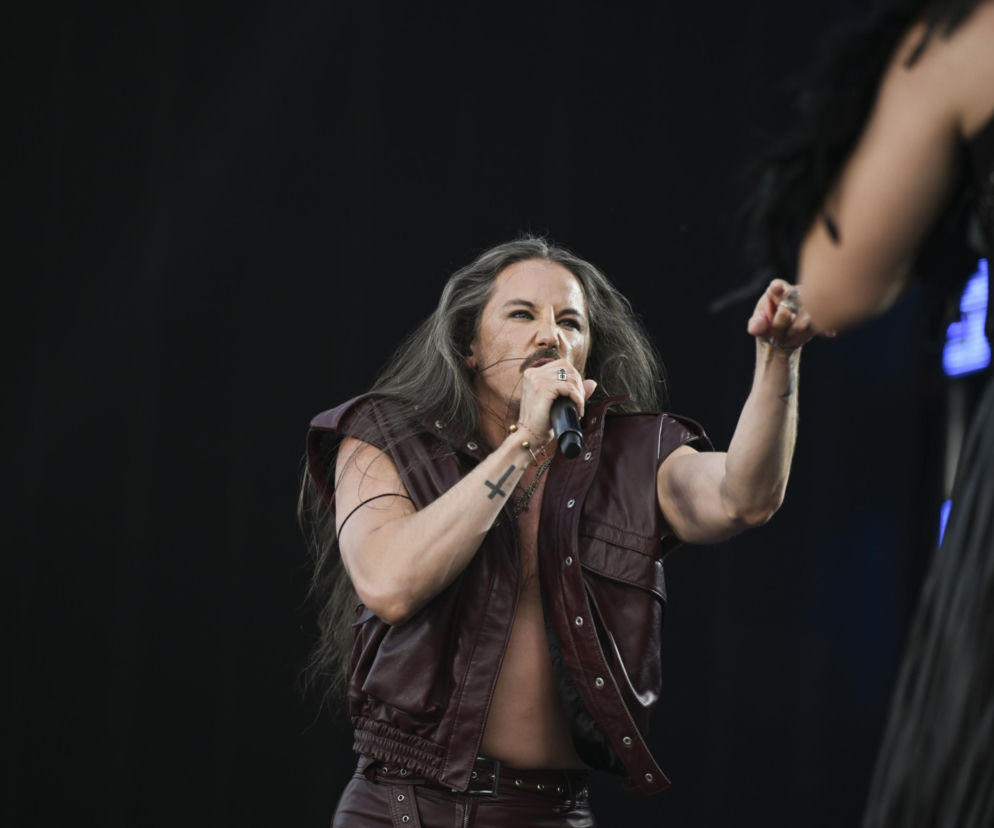 Wokalista Michał Szpak w skórzanej, bordowej kamizelce i spodniach z długimi, siwymi włosami i charakterystycznym wąsem, trzymający mikrofon podczas występu. Artysta, znany ze swojego wizerunku scenicznego, opowiedział o swoim lęku, o czym przeczytasz na naszym portalu.