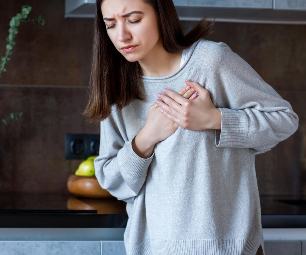 Kobieta w jasnoszarym swetrze trzymająca dłonie na klatce piersiowej, z zamkniętymi oczami i zmartwionym wyrazem twarzy, odczuwająca ból w sercu lub kołatanie po posiłku. Tło stanowi fragment nowoczesnej kuchni z ciemnymi kaflami i zieloną rośliną w wazonie. Dowiedz się więcej o przyczynach i objawach kołatania serca na Poradnik Zdrowie.