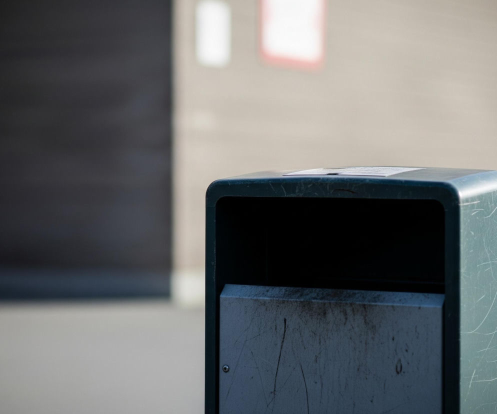 Na pierwszym planie, z prawej strony kadru, znajduje się ciemnozielony metalowy kosz na śmieci o zaokrąglonych narożnikach. Jego powierzchnia jest mocno porysowana, a na wewnętrznej, jaśniejszej klapie widoczne są ślady brudu i zacieków. Na górnej części kosza leży biała kartka papieru. Tło jest rozmyte, z lewej strony widnieje ciemnoszara, prostokątna powierzchnia, a za koszem jasnobrązowa ściana z prostokątnym, rozświetlonym oknem lub tablicą z czerwoną obwódką w oddali.