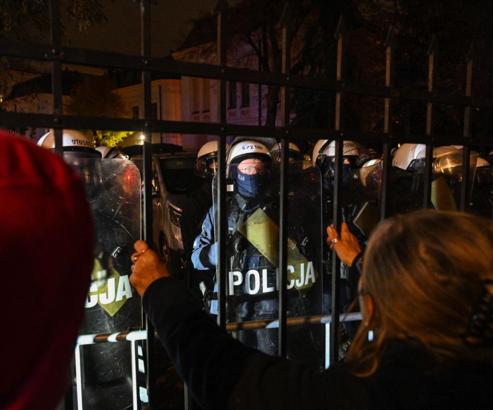Kobieta trzyma ręce na metalowym płocie. Za nim policjanci z tarczami. O protestach przed TK przeczytasz w serwisie Eska.