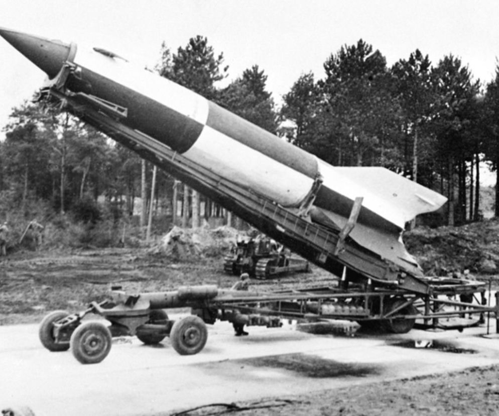 Czarno-białe zdjęcie rakiety balistycznej V-2 w pozycji startowej na ruchomej wyrzutni w lesistym terenie. Obiekt, znany jako cudowna broń Hitlera, został przejęty przez Armię Krajową na Podlasiu. Więcej o operacji Most III i jej znaczeniu dla losów II wojny światowej przeczytasz na naszym portalu.
