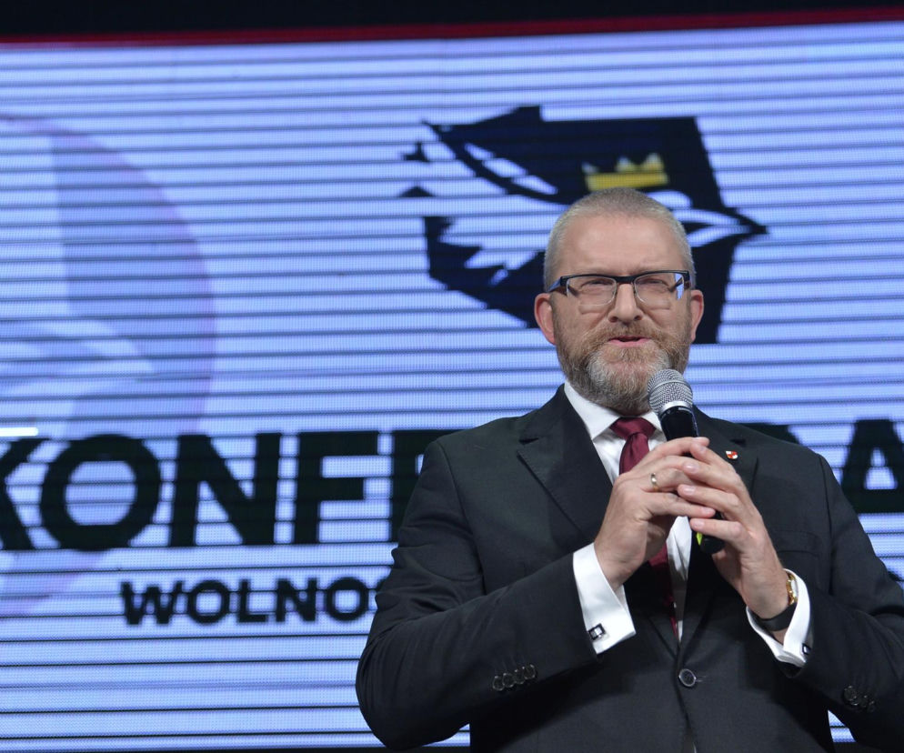 Grzegorz Braun w garniturze przemawia do mikrofonu na tle logo Konfederacji. O jego procesie czytaj na SE Polityka.