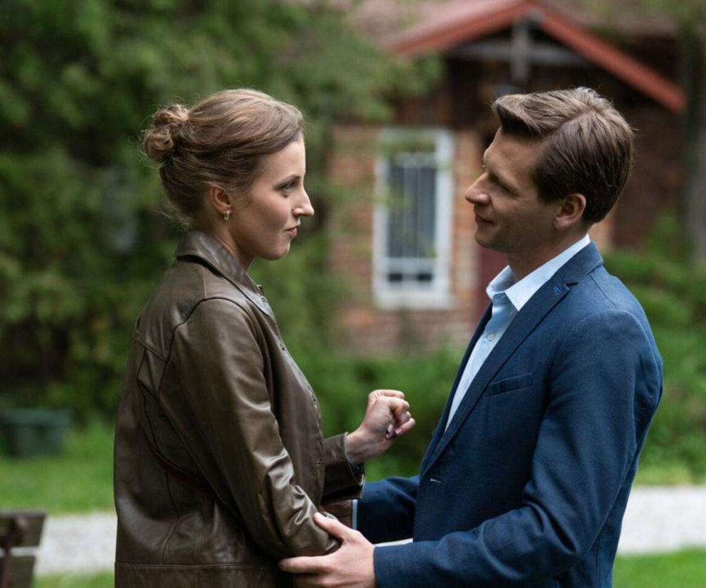 Natalka (Dominika Suchecka) i Adam Karski (Patryk Szwichtenberg) z M jak miłość stoją naprzeciwko siebie w ogrodzie, trzymając się za ręce. Natalka ma zamyśloną minę, a Adam patrzy na nią z nadzieją. W tle widać zieleń i fragment domu. Więcej o ich losach na Super Seriale.