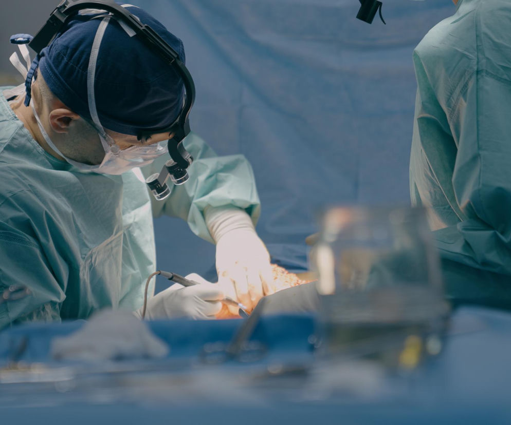 Dwóch chirurgów w zielonych fartuchach operacyjnych, w czepkach i z lupami operacyjnymi na głowach, pochylonych nad otwartą klatką piersiową pacjenta, wykonujących skomplikowaną operację przeszczepu płuc. Artykuł o tym, jak lekarze usunęli oba płuca 33-latkowi, dostępny jest na portalu Poradnik Zdrowie.