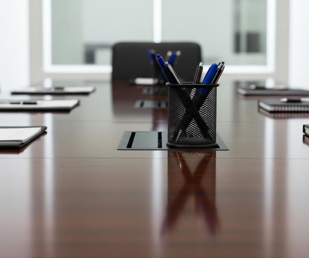 Centralnym elementem obrazu jest koszyk na długopisy, wykonany z czarnej, metalowej siatki, zawierający kilka czarnych i niebieskich długopisów, stojący na wypolerowanym, brązowym stole konferencyjnym. Stół odbija światło i obiekty, w tym koszyk i widoczne na nim elementy. Na stole po obu stronach koszyka leżą otwarte foldery lub podkładki z białymi kartkami papieru oraz zamknięte notatniki z czarnymi okładkami i spiralnymi grzbietami, a na każdym z nich spoczywa jeden długopis. W tle znajdują się czarne, tapicerowane krzesła, a za nimi widać jasne, białe ściany oraz okna, przez które prześwituje rozmyte światło.