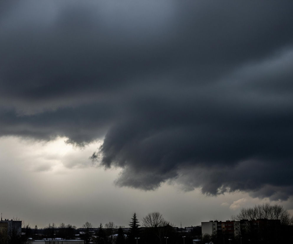 Duże, ciemnoszare chmury dominują niebo, rozciągając się horyzontalnie na większości kadru. Poniżej nich, w środkowej części obrazu, widoczne jest jaśniejsze, szare niebo. Na dole zdjęcia, ciemne sylwetki budynków mieszkalnych o płaskich dachach i nagich drzew, a także kilka mniejszych, iglastych drzew, tworzą horyzontalny pas. Fragmenty śniegu leżą na ziemi między budynkami i drzewami, a gdzieniegdzie widać ciemne latarnie uliczne.