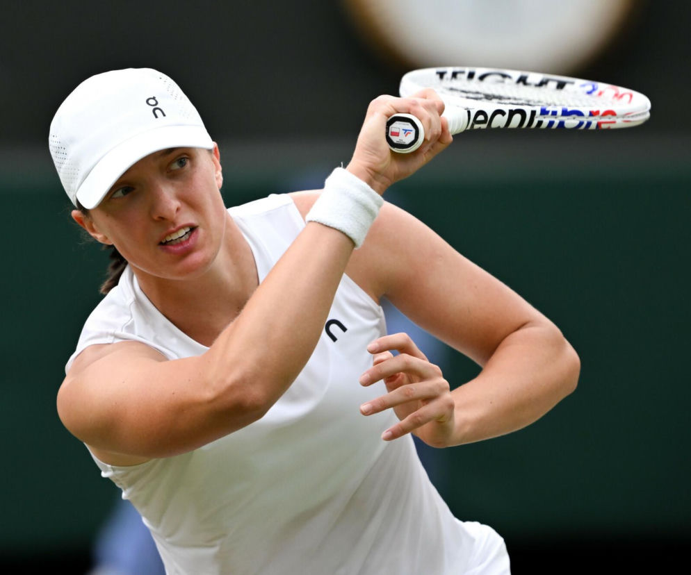 Iga Świątek, polska tenisistka, w białej czapce i sportowym stroju, przygotowuje się do uderzenia rakietą podczas meczu na korcie Wimbledonu. Jej skupiona mimika i dynamiczna pozycja ciała oddają intensywność sportowej rywalizacji. Więcej o relacji na żywo przeczytasz na naszym portalu.