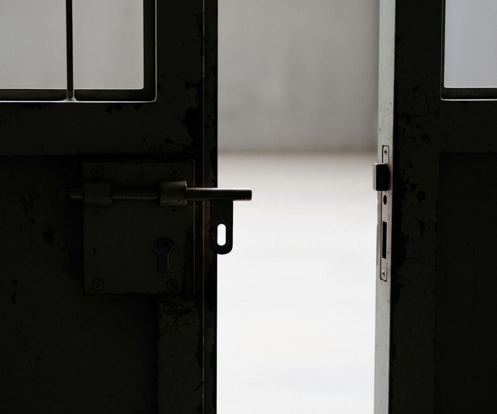 Częściowo otwarte, ciemne, metalowe drzwi z kratami, lekko uchylone w kierunku jasnego, rozmytego tła. Po lewej stronie widoczna jest skrzydło drzwi z wystającym ryglem i zamkniętym mechanizmem zamka. Górna część drzwi zawiera kraty, przez które przebija się światło z zewnątrz, tworząc na powierzchniach metalu refleksy i odcienie szarości, kontrastując z niemal czarną dolną częścią drzwi, pełną tekstury i śladów zużycia. Pomiędzy skrzydłami drzwi widać wąski, pionowy pas jasnego światła.