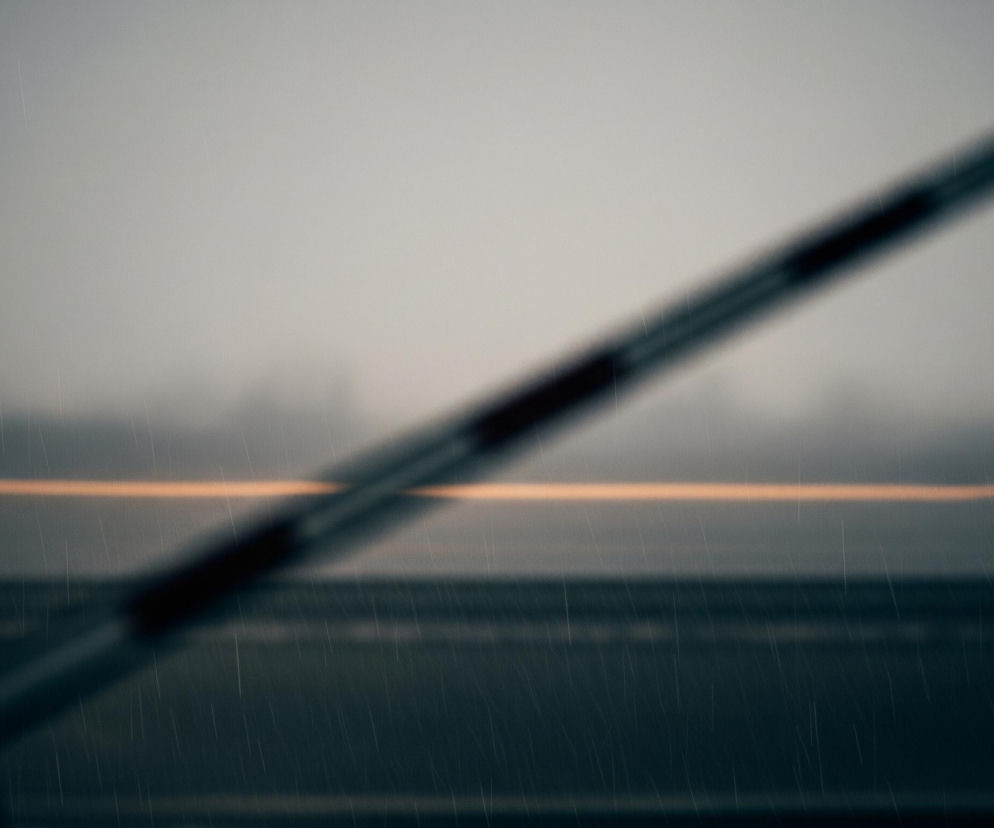 Główny obiekt to rozmyty, podłużny przedmiot w ukośnej pozycji, przechodzący przez centrum kadru. Jest on ciemny, z widocznymi jaśniejszymi, poziomymi pasami. W tle widoczna jest szeroka, ciemnoszara płaszczyzna, przedzielona w połowie horyzontalną, pomarańczową linią. Cały obraz pokryty jest efektem deszczu, widocznym jako drobne, jasne smugi.